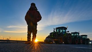 Landwirte blockieren am frühen Morgen im brandenburgischen Jacobsdorf mit ihren Traktoren die Auffahrt auf die Autobahn A12 in Richtung Berlin, ein Mann steht dabei im Gegenlicht der aufgehenden Sonne.