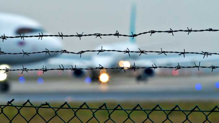 Hinter einem Zaun mit Stacheldraht sind Flugzeuge auf dem Rollfeld eines Flughafens zu sehen.