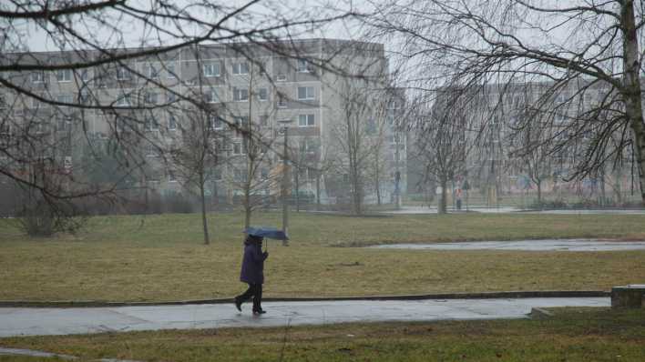 Eine Frau geht im stroemenden Regen durch einen Park in Brandenburg an der Havel, im Hintergrund unsanierte Plattenbauten.(Bild: picture alliance / Caro / Hechtenberg)