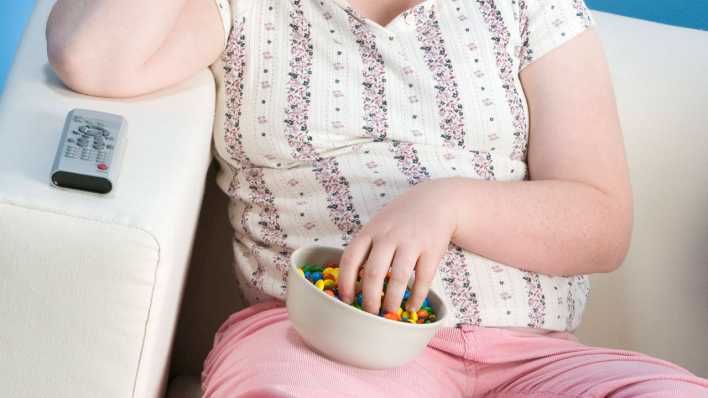Übergewichtiges Mädchen sitzt mit Süßigkeiten vor dem Fernseher