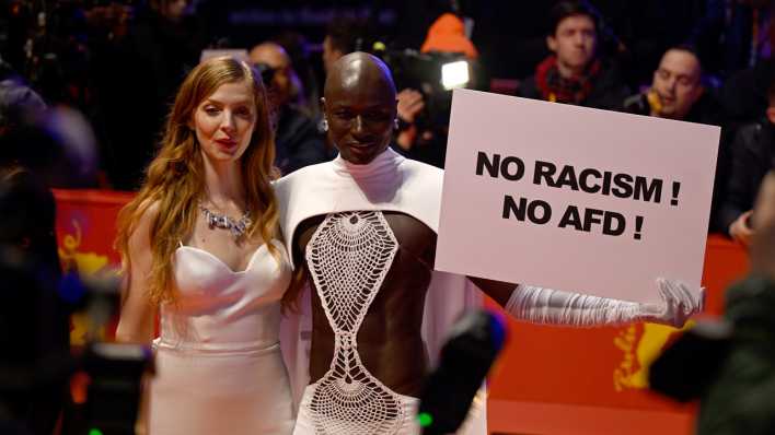 Papis Loveday (r) steht am Eröffnungsabend der Berlinale mit einem Protestschild "No Racism ! No AFD !" neben Schauspielerin Pheline Roggan auf dem Roten Teppich. (Bild: Monika Skolimowska/dpa)