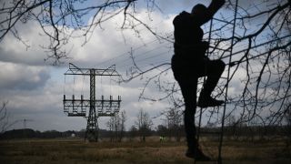 Ein Journalist klettert auf einen Hochsitz, während die Polizei an einem beschädigten Strommasten auf einem Feld in der Nähe der Tesla-Autofabrik in Grünheide ermittelt (Bild: picture alliance/dpa/Sebastian Gollnow)