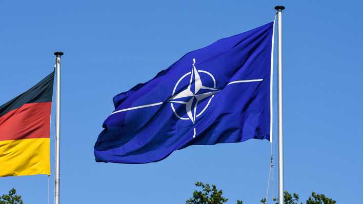 Die wehende Flagge der NATO und im Anschnitt eine Deutschlandflagge an der Zufahrt zum Fliegerhorst.