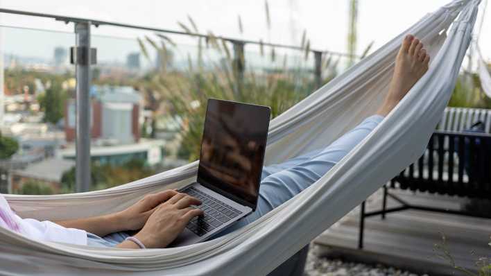 Eine jung Frau liegt in einer Hängematte und arbeitet am Laptop.