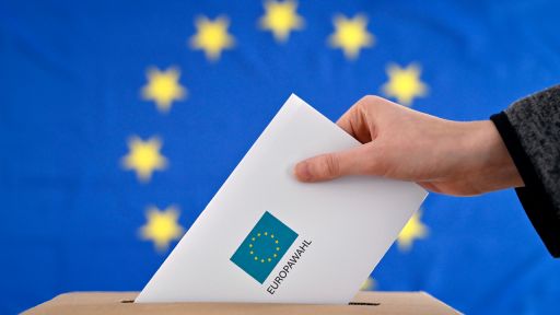 Ein Wahlbrief wird in eine Wahlurne gesteckt, im Hintergrund eine EU-Flagge.