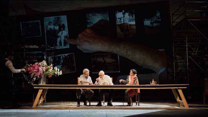 Drei Menschen sitzen an einem langen Tisch in einer Szene aus dem Theaterstück "House".
