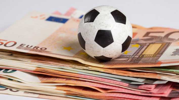 Ein Fußball liegt auf einem Stapel mit Geld und repräsentiert die Verflechtung von Spielergebnissen und finanziellen Auswirkungen.