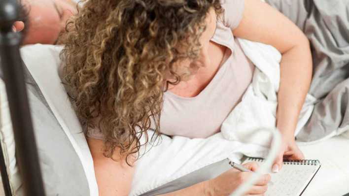 Frau liegt in einem Bett und notiert sich etwas in einem Notizblock (Bild: picture alliance/dpa Themendienst/Christin Klose)