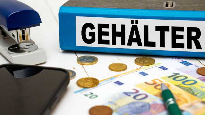 Gehälter steht auf einem blauen Aktenordner neben Geld-Scheinen und Euro-Münzen auf einem Schreibtisch (Bild: picture alliance/CHROMORANGE/Michael Bihlmayer)
