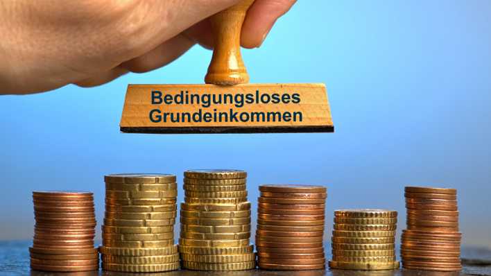 Symbolbild: Holzstempel mit Aufschrift "Bedingungsloses Grundeinkommen", gehalten von einer Hand im Anschnitt über mehreren Stapeln mit Euro-/Cent-Münzen (Bild: picture alliance / SULUPRESS.DE / Torsten Sukrow)