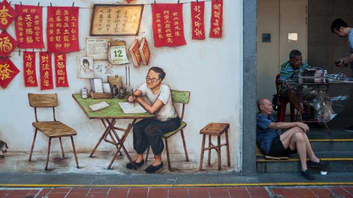 Ein älterer Mann sitzt auf einer Treppenstufe neben einem Wandbild in Singapur.