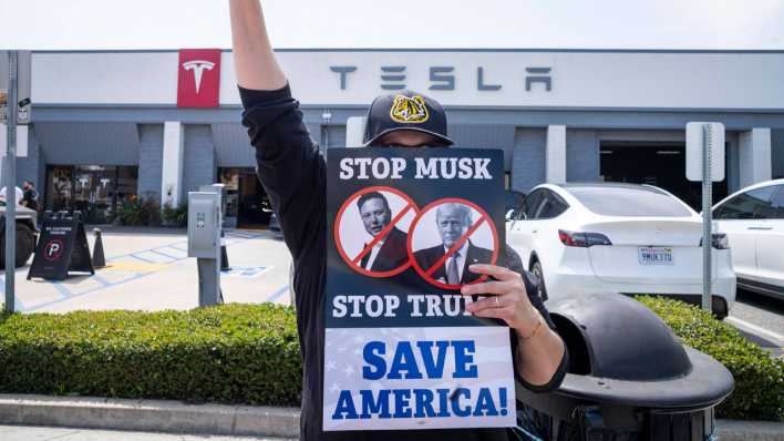 Ein Demonstrant hält vor einer Tesla-Filiale ein Schild hoch mit der Aufschrift "Stop Musk, Stop Trump - Save America".