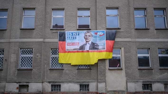 Ein Wahlplakat von Hans-Christoph Berndt, Vorsitzender der Brandenburger AfD-Fraktion, hängt auf einer Deutschlandflagge an einer tristen Hausfassade.