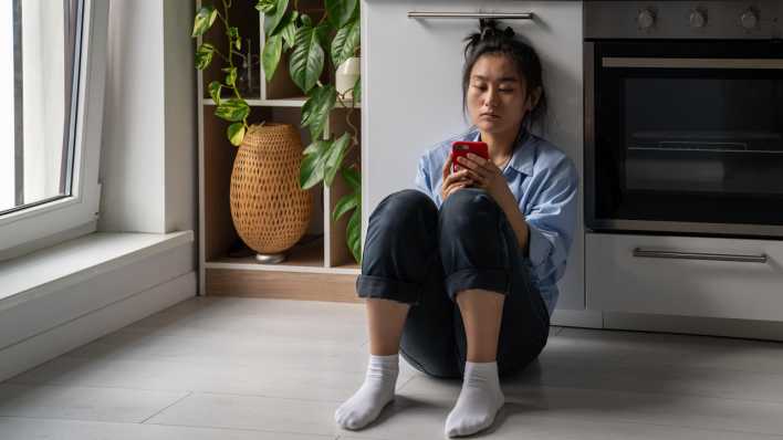 Eine traurige Frau sitzt auf dem Boden und schaut aufs Smartphone. (Bild: picture alliance / Zoonar | Dmitrii Marchenko)
