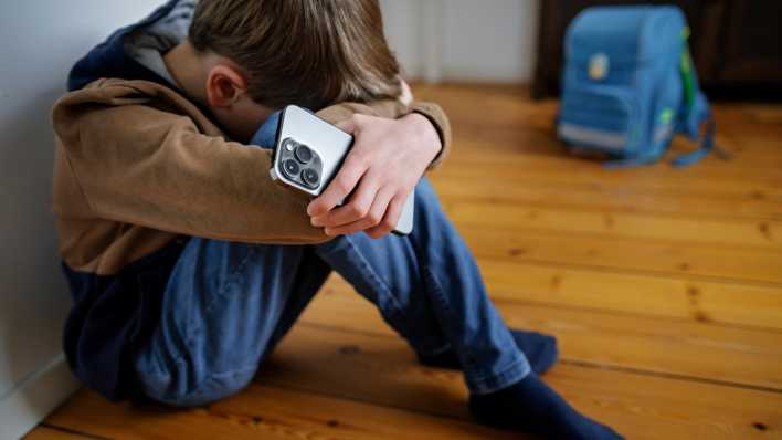 Ein Junge sitzt mit einem Smartphone in der Hand neben seinem Schulranzen und versteckt seinen Kopf.