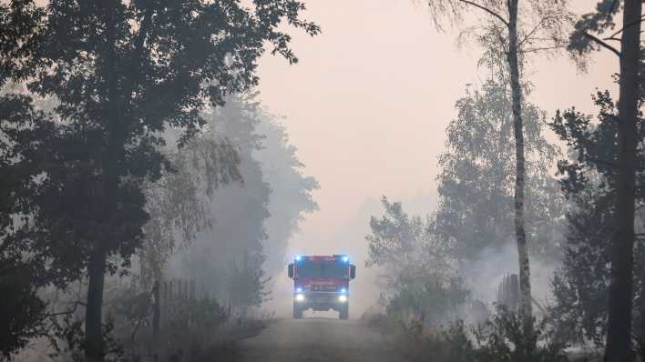 Ein Feuerwehrfahrzeug fährt am 26.07.2022 über einen verrauchten Weg während eines Waldbrandes bei Falkenberg. (Quelle: dpa/Jan Woitas)